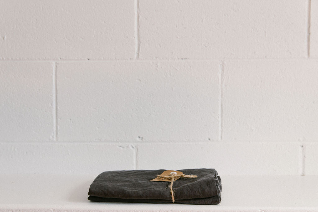 A French linen duvet cover Charcoal colour on a white brick background, neatly folded and tied with brown twine and a brown kraft paper tag.
