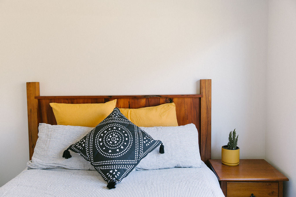 Bed with wooden headboard, yellow pillows with black and white throw cushion by kim soo, and a small plant on a nightstand.