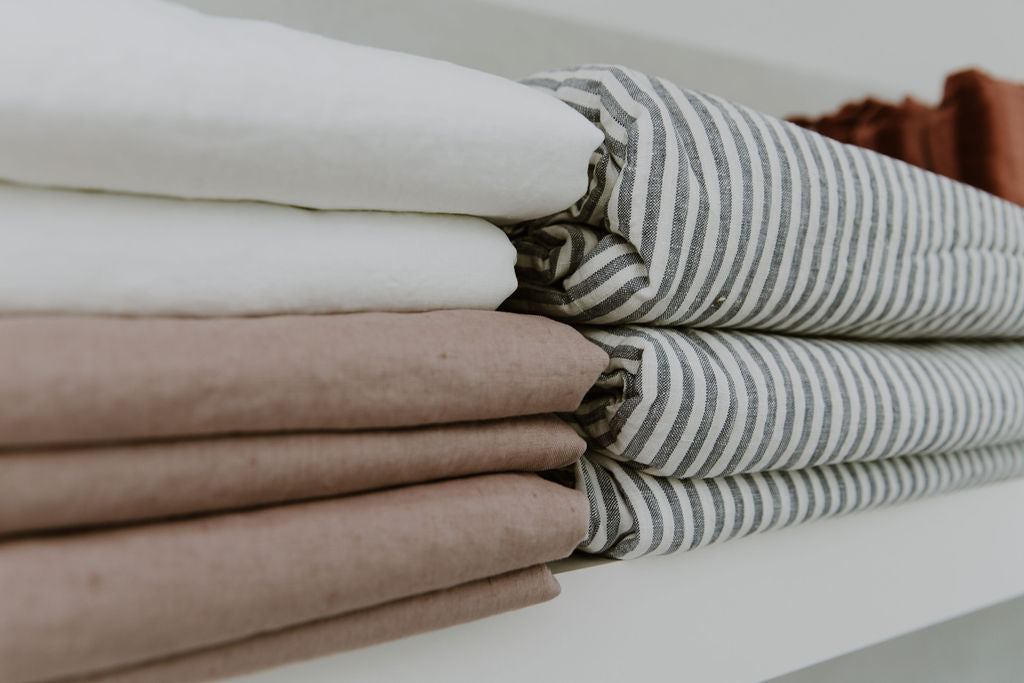 Stack of folded French linen sheets in crisp white, blush and marine stripe colours on a light surface.