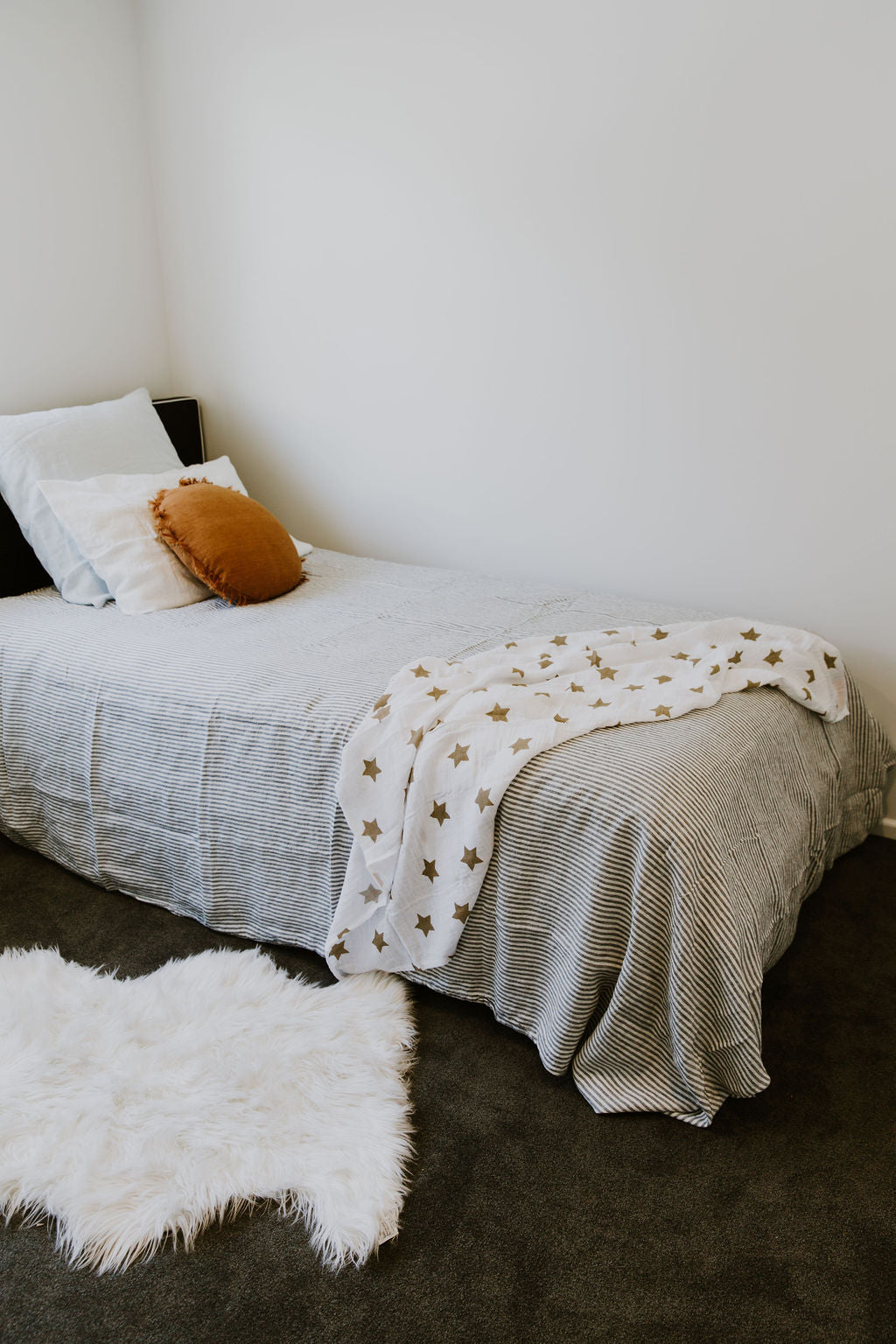 Neatly made bed with grey striped French linen bedding and a white blanket with gold stars, surrounded by a white rug on a dark floor.