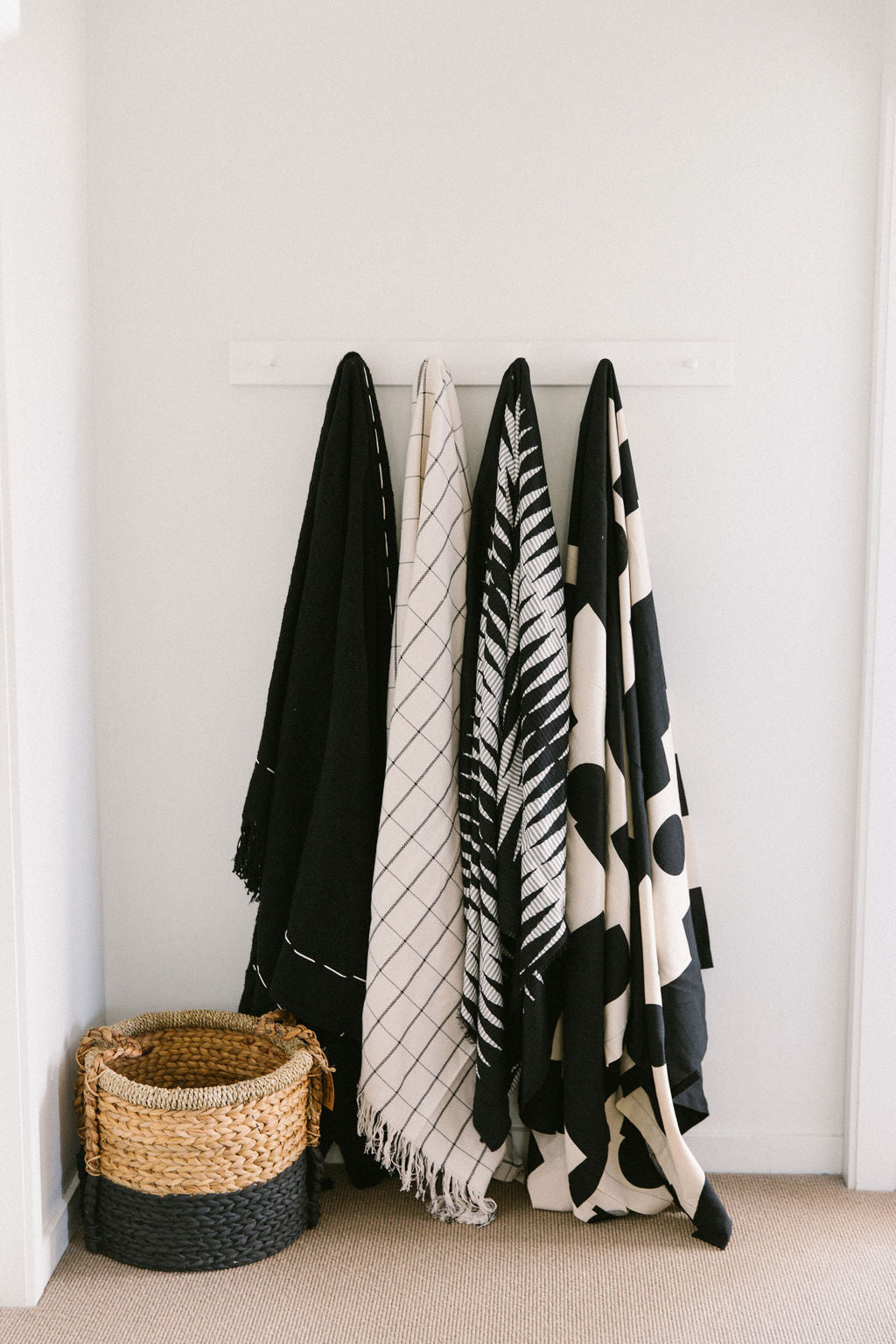 Black, white, and patterned towels hanging on a rack against a white wall with a seagrass basket on the ground beside