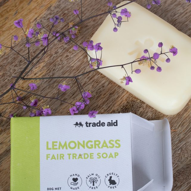 Lemongrass fair trade soap bar with packaging on a wooden surface with purple flowers.
