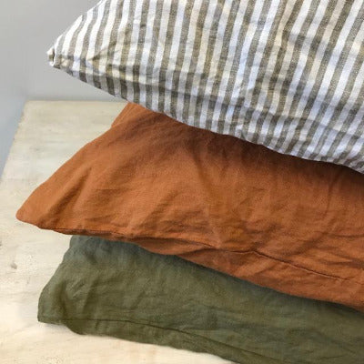 The corners of three pillows made from French flax linen, in olive stripe, burnt orange, and olive colours. Stacked on a light wooden table with a white background.
