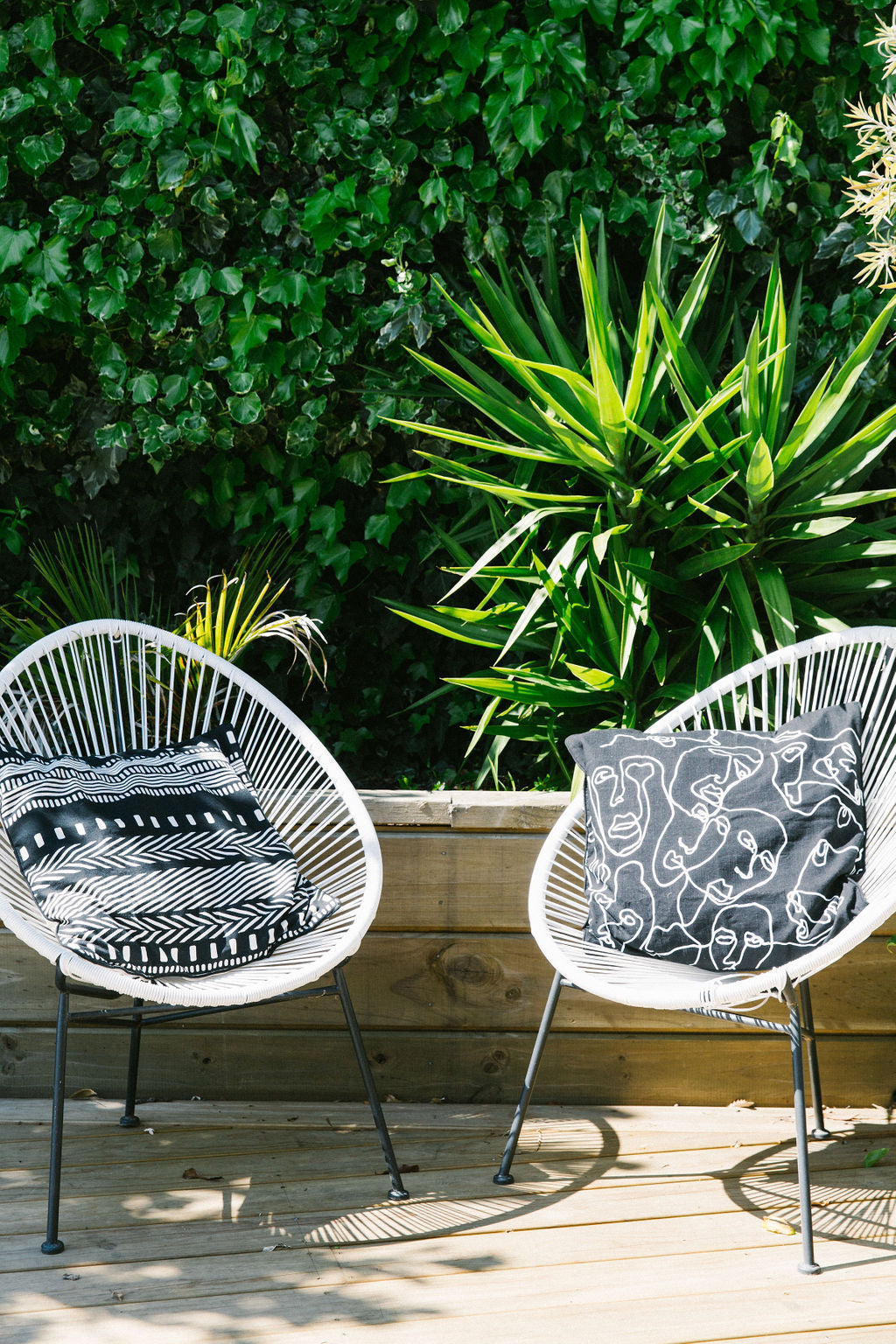 Two white wire chairs with kim soo black and white patterned cushions on a wooden deck surrounded by greenery.