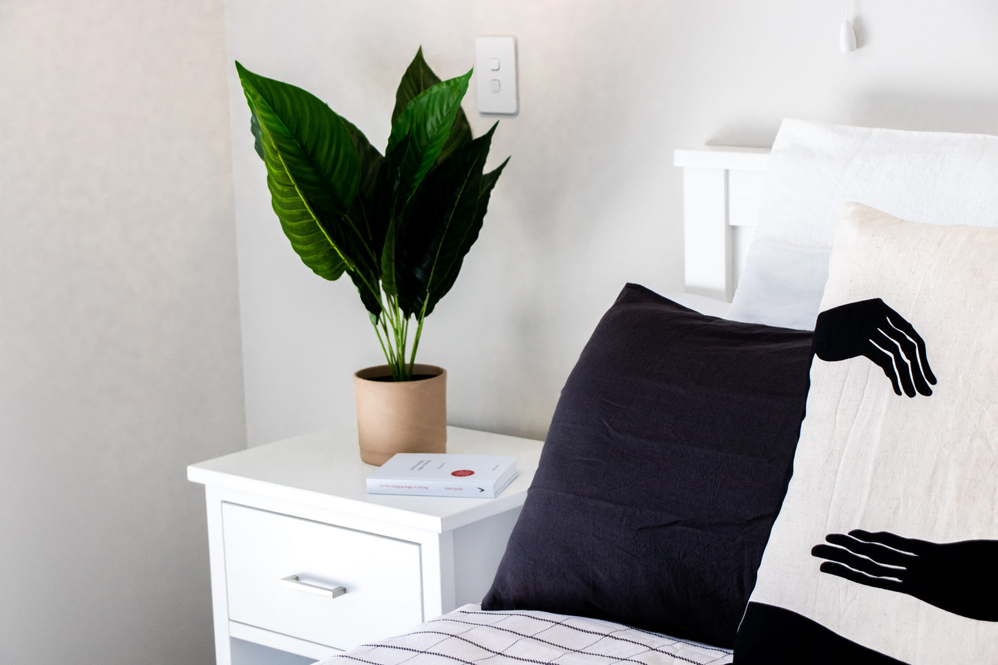 Bedroom with a plant on a nightstand and a black pillow, with a close up of a kim soo linen rayon pillow, with black hands on a natural background