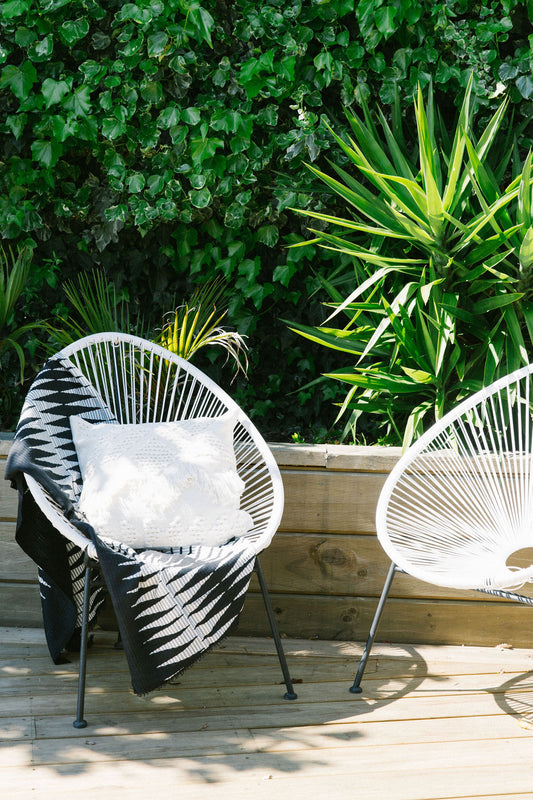 Two outdoor chairs on a wooden deck, a balinese throw and decorative cushion, with greenery in the background