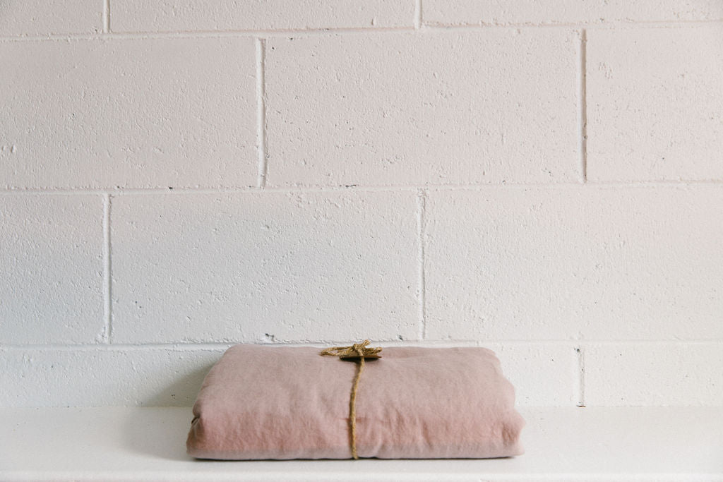 A French linen duvet cover blush colour on a white brick background, neatly folded and tied with brown twine and a brown kraft paper tag. 