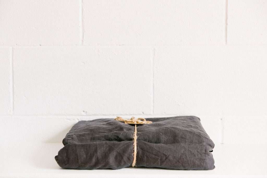 A neatly folded charcoal coloured French flax linen flat sheet, tied with brown twine with a white brick background.
