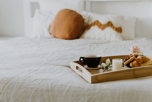Breakfast tray with coffee and pastries on a bed made neatly with French linen in a crisp white colour and decorative cushions in white and burnt orange colours. 