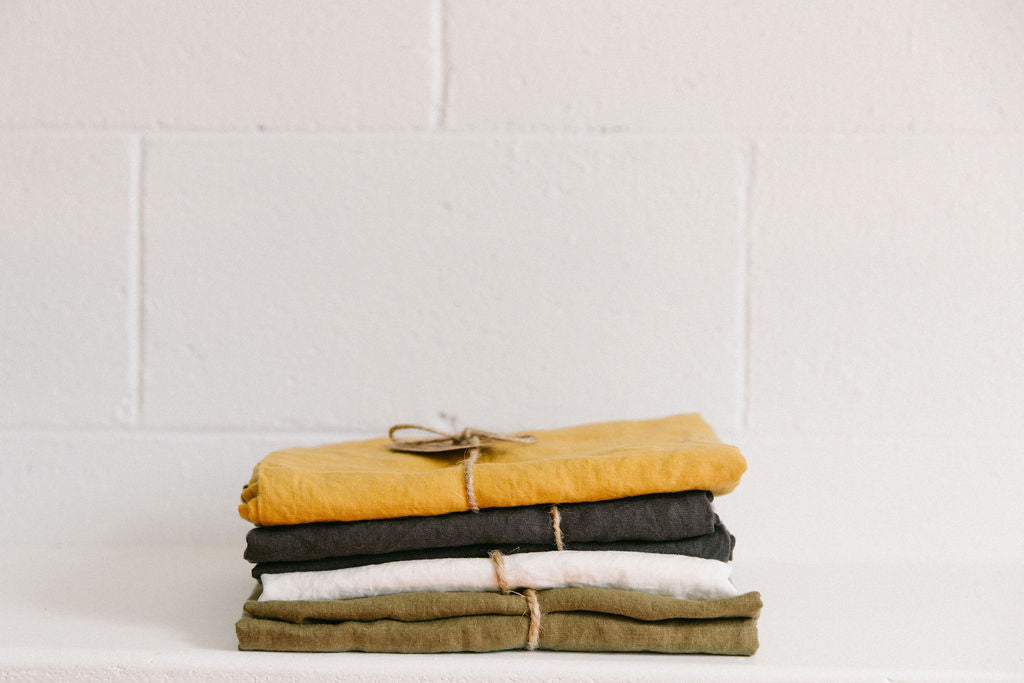 Stack of folded French flax linen on a white surface with a white brick wall background. Colours, Mustard, Charcoal, Crisp White and Olive