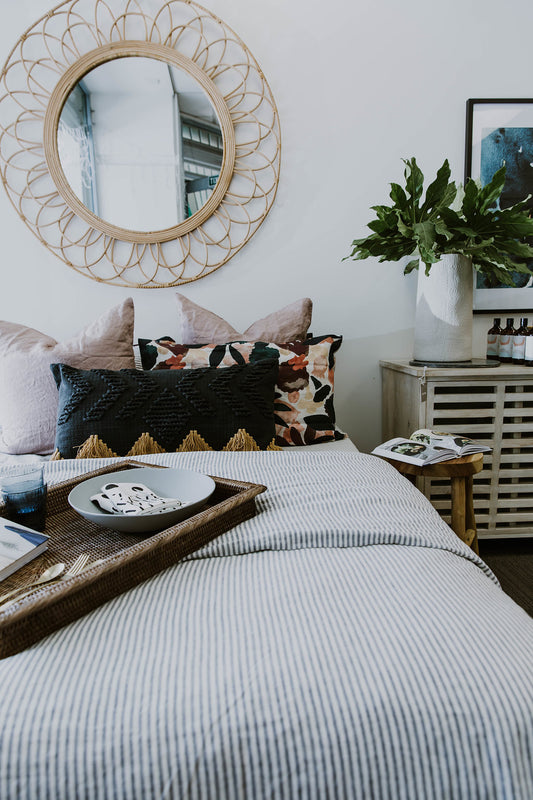 Decorative bedroom with round mirror, pillows, and a plant, on a French linen duvet with navy and off white stripes.