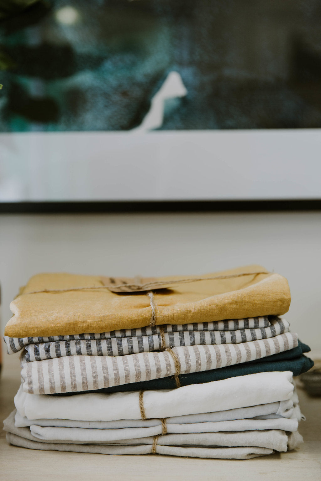 Stack of folded French linen sheets including mustard, navy stripe, grey stripe, crisp white and grey. Tied individually with brown twine and a kraft paper tag on a light coloured table. Blurred framed picture in the background with dark greenery. 