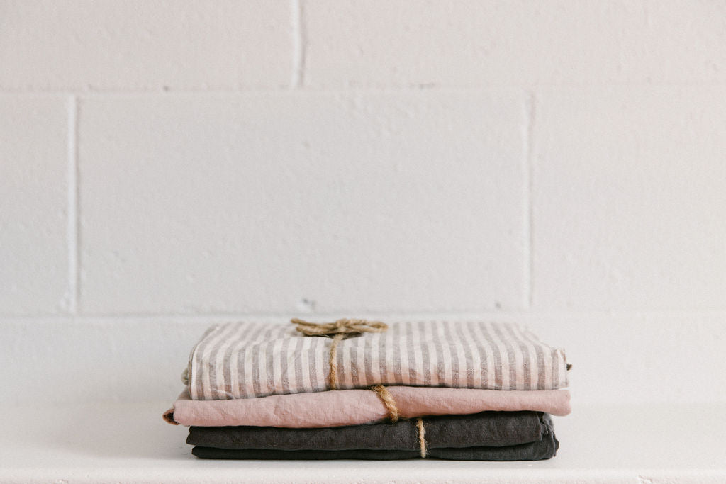 Stack of folded linen sheets including grey stripe, blush and charcoal colours on a white surface in front of a white brick wall. Linen separately tied with brown twine