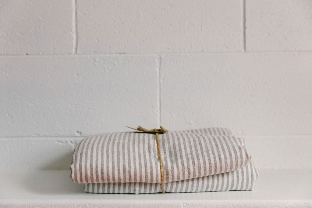 A folded over duvet cover in a grey and white striped colour, made of French flax linen, displayed on a flat white surface with a white brick background.