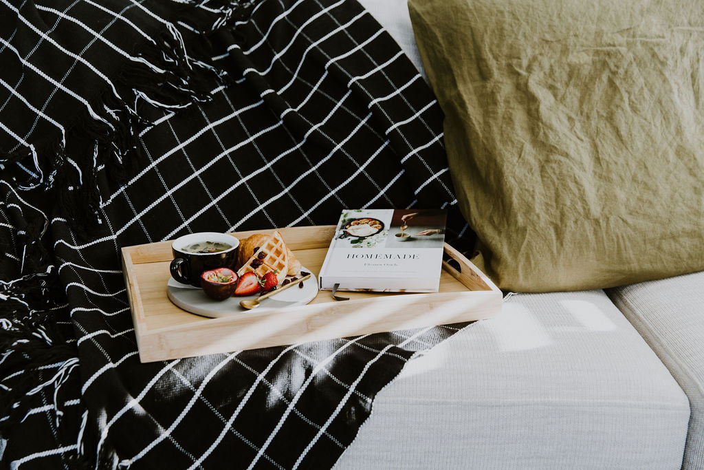 Wooden tray with food and a book on a bed with black and white checkered throw blanket and olive french linen pillow.