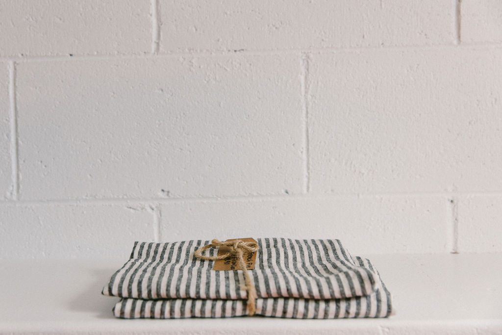 A French linen duvet cover in a navy and off white marine stripe colour on a white brick background, neatly folded and tied with brown twine and a brown kraft paper tag. 