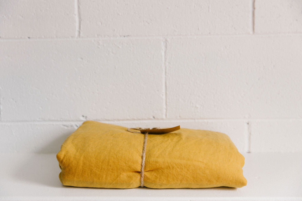 A French linen duvet cover mustard colour on a white brick background, neatly folded and tied with brown twine and a brown kraft paper tag. 