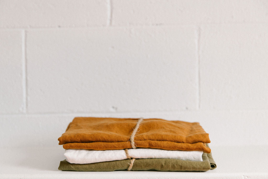 Stack of neatly folded French flax linen sheets including burnt orange, crisp white and olive colours. Tied individually with brown twine on a light surface with a white brick background.
