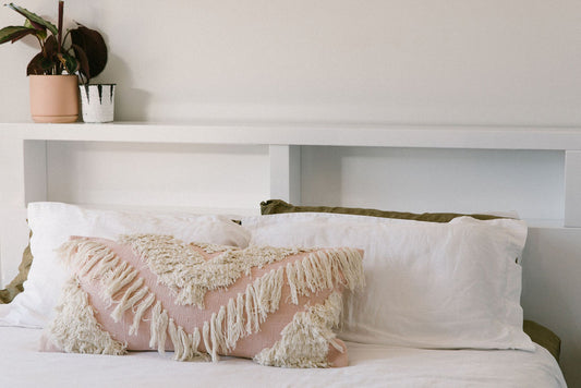 Bed with white pillows and a decorative pink pillow with fringe, against a neutral wall.