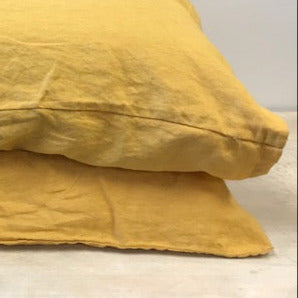 The corner of two pillows with mustard yellow coloured French flax linen pillowcases, stacked on a light wooden table with a white background. 