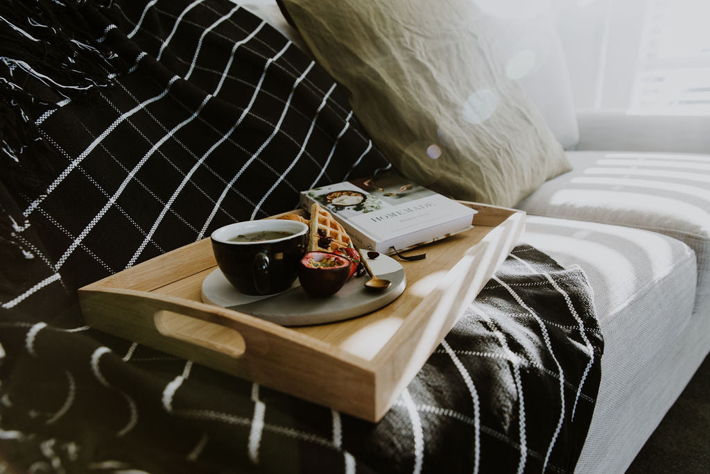 Wooden tray with a cup, saucer, and small plate on a couch with a hand loomed cotton throw blanket and pillow.