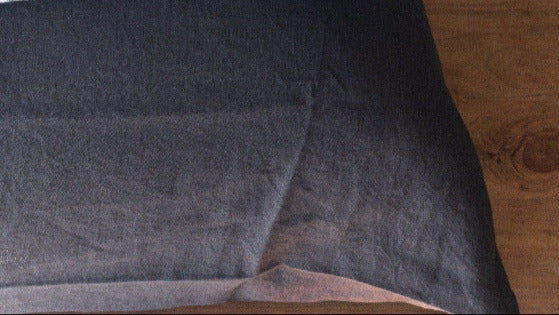 The corner of a pillow with a charcoal coloured French flax linen pillowcase, on a wooden surface